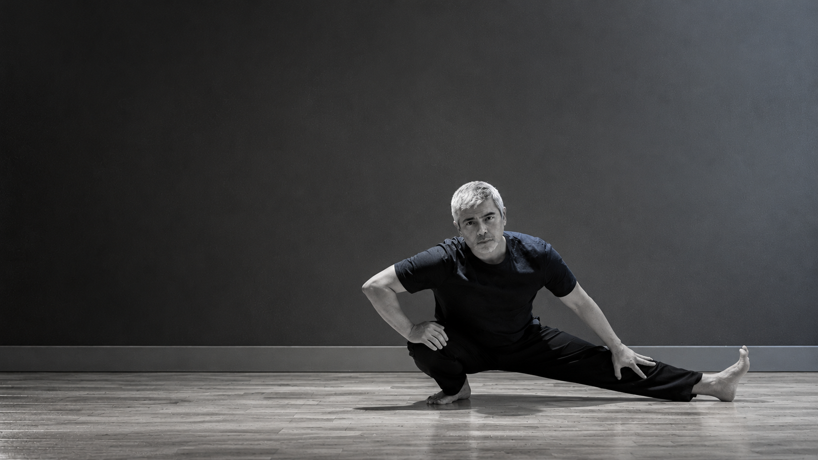 Eugene Salleh holding a Cossack squat on the right of a black-and-white studio photograph